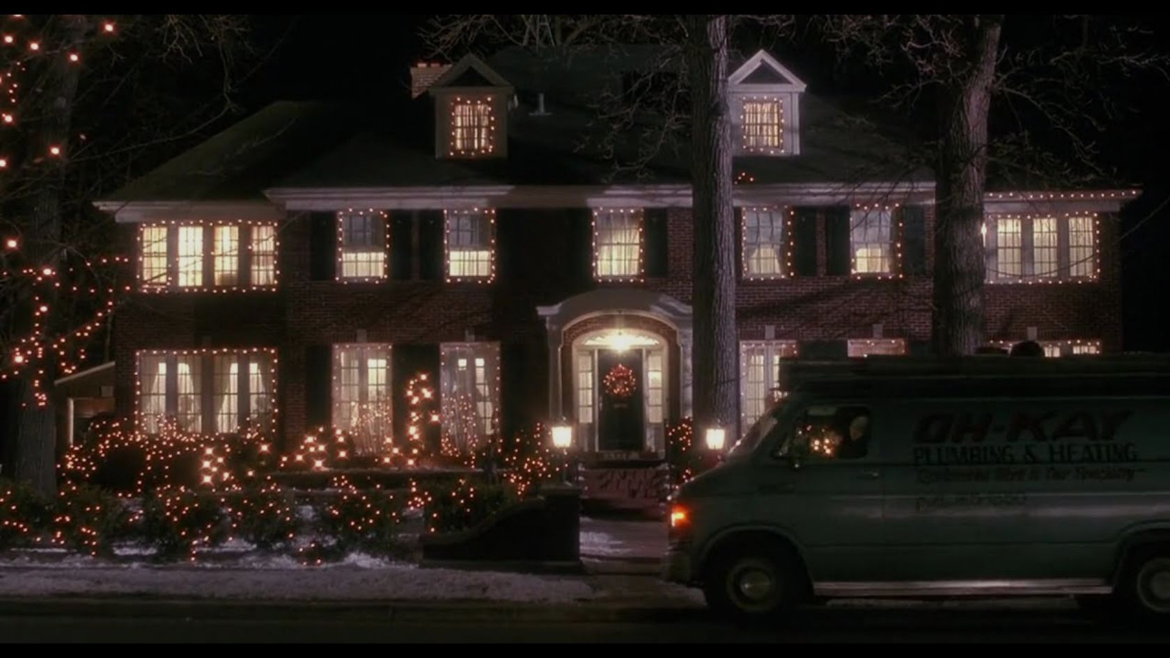 Two-story brick house at night adorned with warm white Christmas lights on windows, trees, and shrubs; a service van is parked in front.
