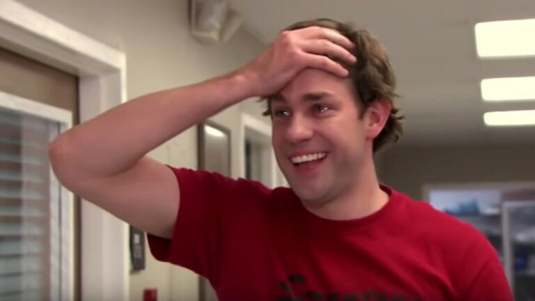Smiling man in a red shirt, hand resting on his forehead in a playful, casual indoor setting.