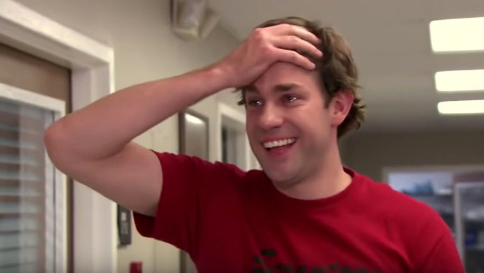 Smiling man in a red shirt, hand resting on his forehead in a playful, casual indoor setting.