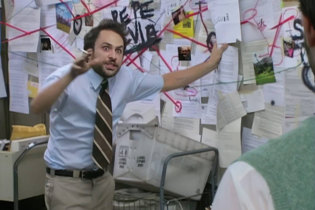 Man in a light blue shirt and striped tie gesturing angrily in a cluttered office covered with papers and red strings connecting clues on a conspiracy-style wall behind him.