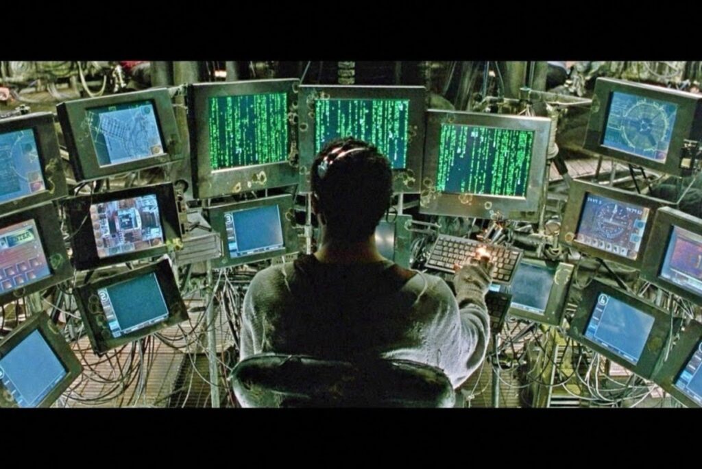 Person seated in a chair at a large, multi-screen computer console with green code on several monitors