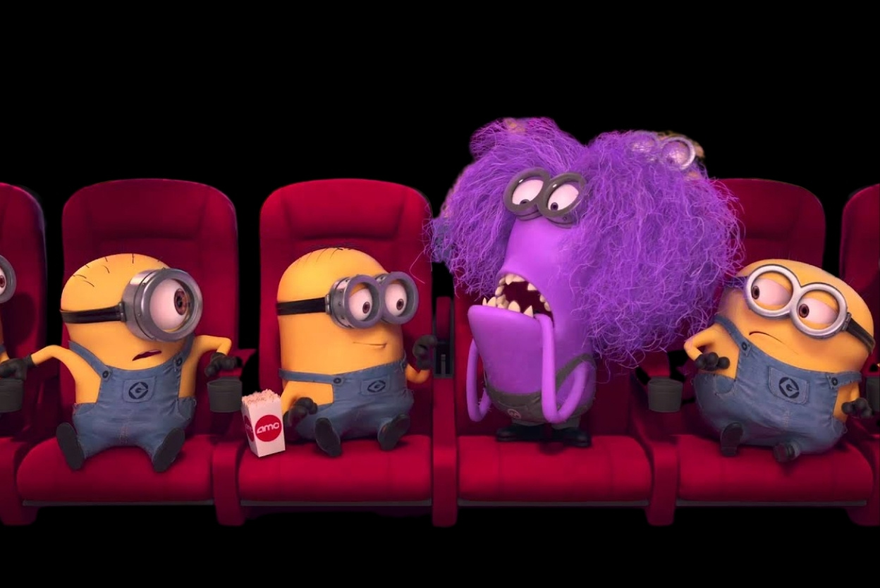 Three Minions in red theater seats with a purple fuzzy monster between them; popcorn nearby, all watching a screen.