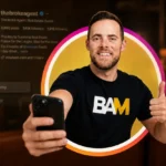 Man taking a selfie with a smartphone, smiling, wearing a black BAM T‑shirt, inside a colorful circular frame behind him.