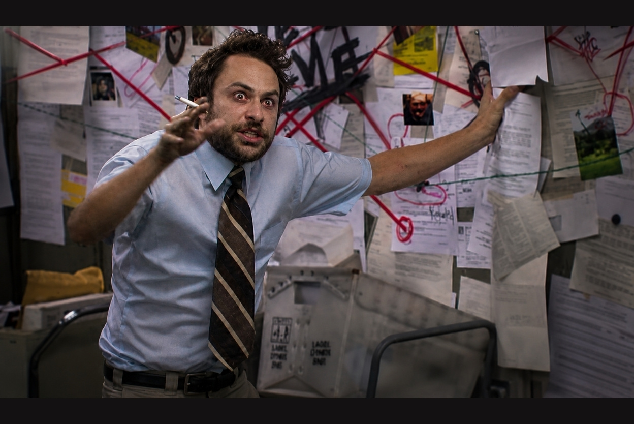 Man in a light blue shirt and striped tie gesticulates aggressively while holding a cigarette, in a cluttered room covered with papers and red string connections behind him.