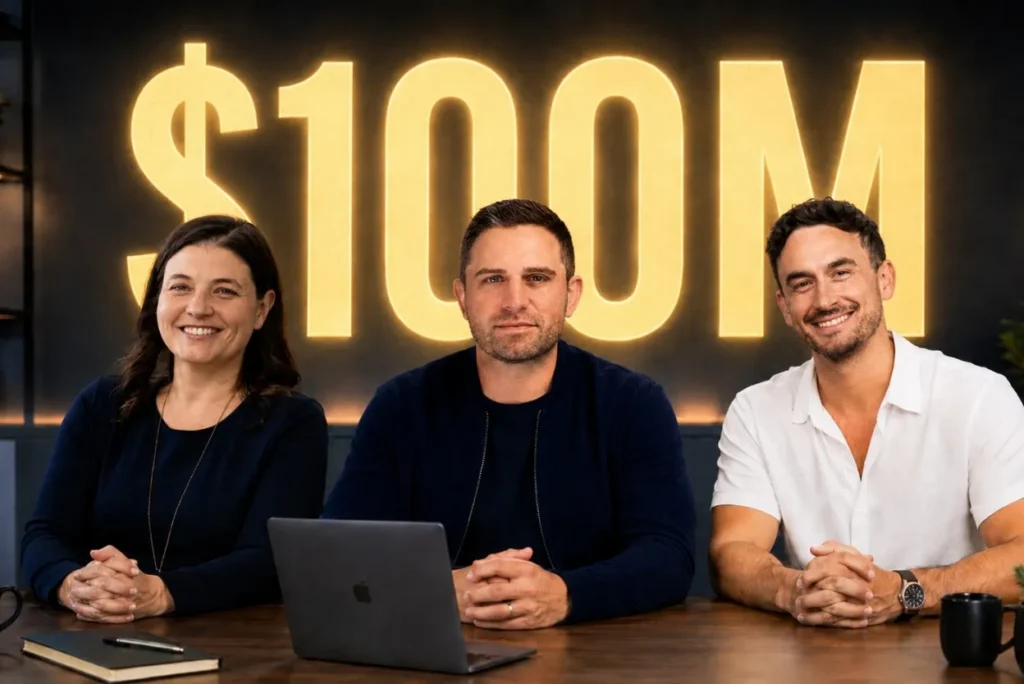 Three people sit at a table in front of a large glowing $100M sign in a studio setting with a laptop and notebook on the table.