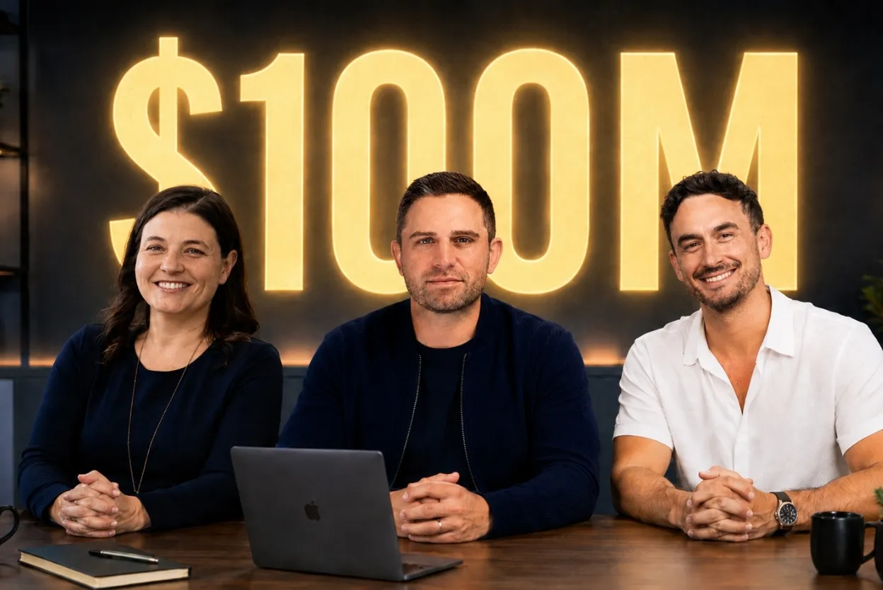 Three people sit at a table in front of a large glowing $100M sign in a studio setting with a laptop and notebook on the table.
