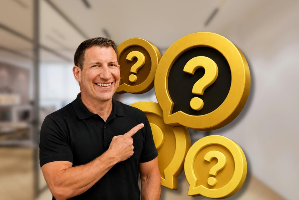 Smiling man in a black polo points to oversized yellow speech bubbles with question marks in an office setting.