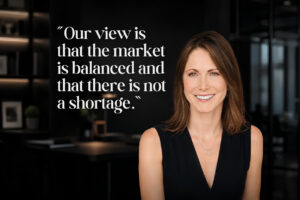 Smiling woman in a black sleeveless top stands in a modern office beside a large white quote about market balance on a dark wall .