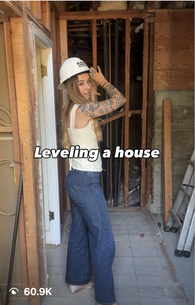 Woman in a white hard hat inside a partially framed house, preparing to level the structure.