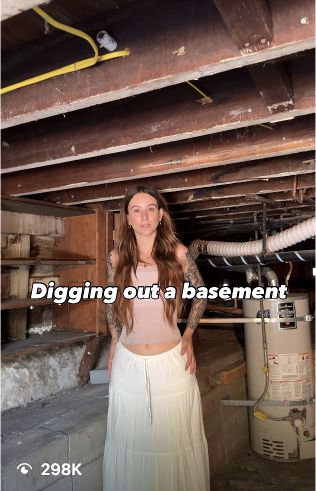 Woman with long hair and tattoos stands in a dim basement under renovation, with exposed wooden joists and plumbing visible behind her.